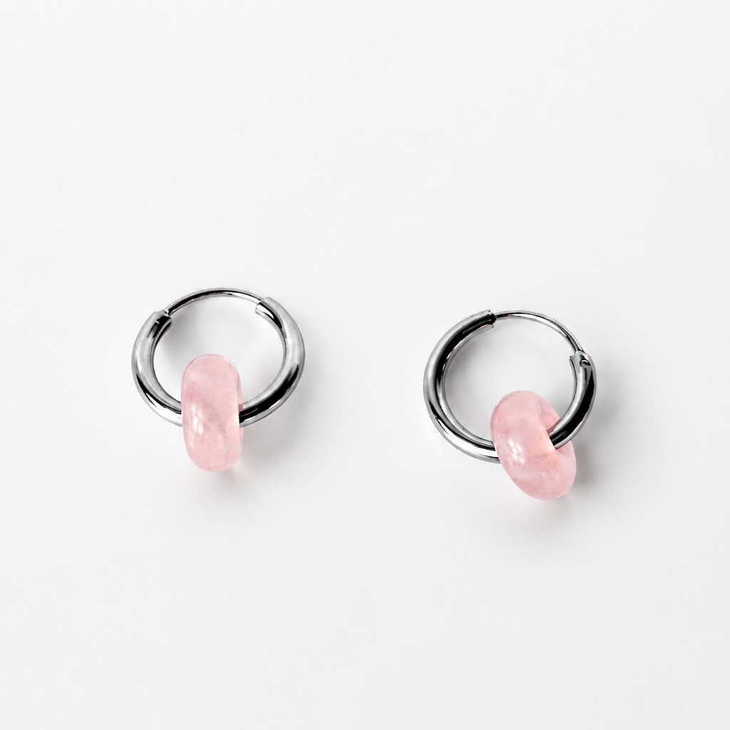 A pair of silver-toned huggie hoop earrings with a small, polished pink stone dangling from the front of each hoop, set against a clean white background.