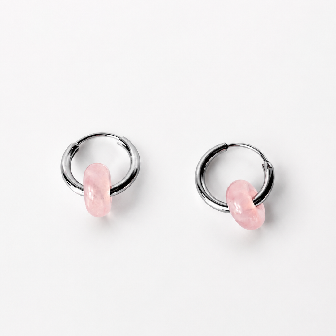 A pair of silver-toned huggie hoop earrings with a small, polished pink stone dangling from the front of each hoop, set against a clean white background.