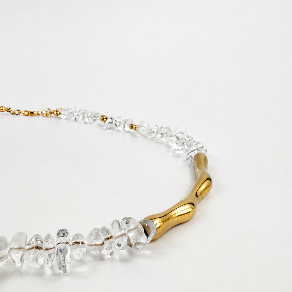 A close-up of a necklace featuring clear faceted crystal-like beads and a polished gold branch-shaped centerpiece on a white background.