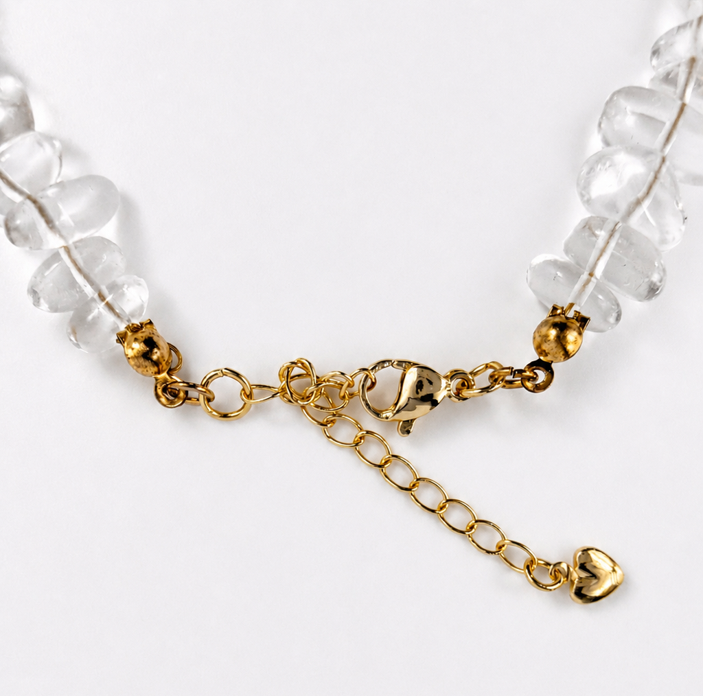 A close-up of a beaded necklace featuring clear faceted stone beads and gold-toned chain details, including a small bell and heart charm, on a plain white background.